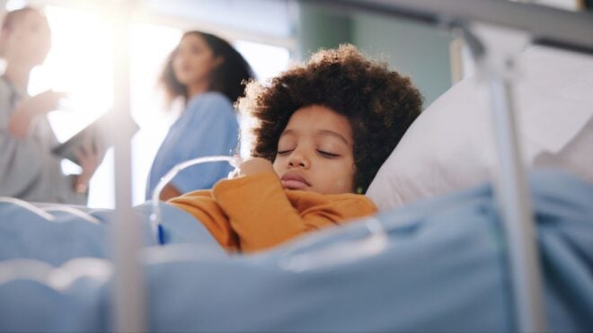 A young child resting in a hospital bed with a parent and doctor having a conversation nearby