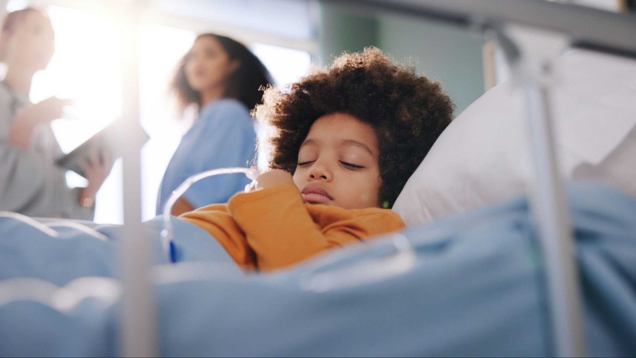 A young child resting in a hospital bed with a parent and doctor having a conversation nearby